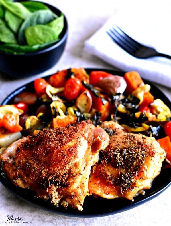 two crispy chicken thighs and roasted vegetables on a black plate with a black fork on a white napkin and a black bowl of spinach leaves.