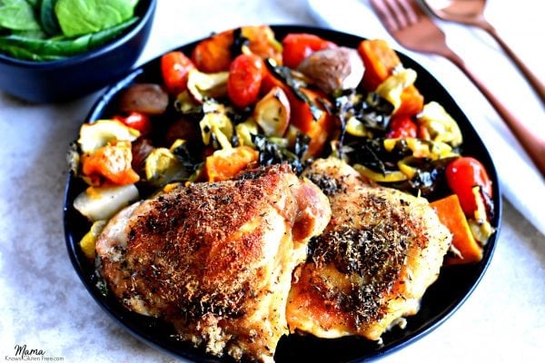 two crispy chicken thighs and roasted vegetables on a black plate with a fork and a black bowl with spinach leaves