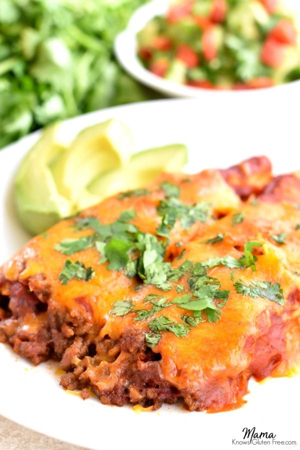 plate of gluten-free beef and cheese enchiladas with sliced avocados and salsa and cilantro in the background