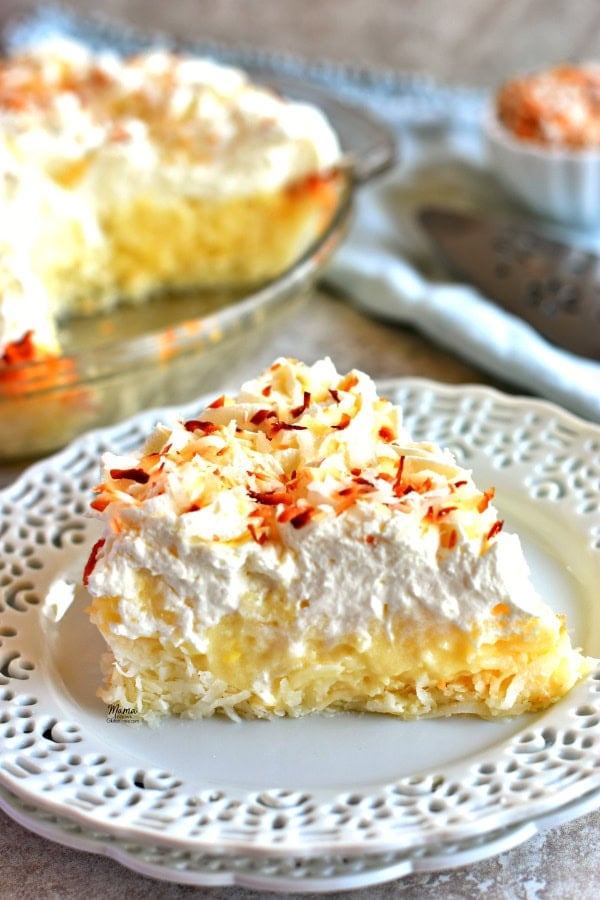 A slice of gluten-free coconut cream pie on a plate with the pie and toasted coconut in the backgorund