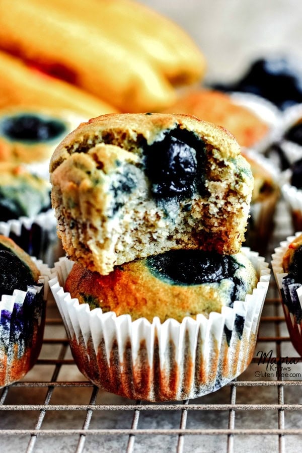 2 Gluten-Free Blueberry Banana Muffins stacked on top of each other with a bite out of one with more muffins and bananas in the background