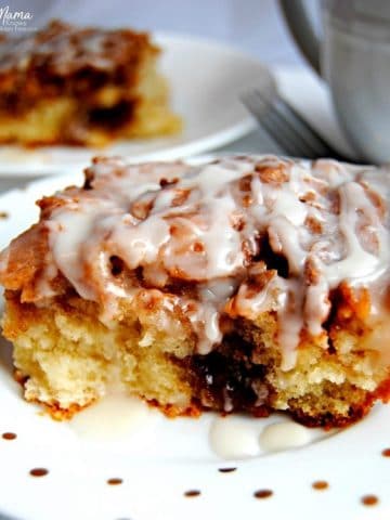 slice of gluten-free cinnamon roll cake on a white plate with another slice and coffee cup in the background