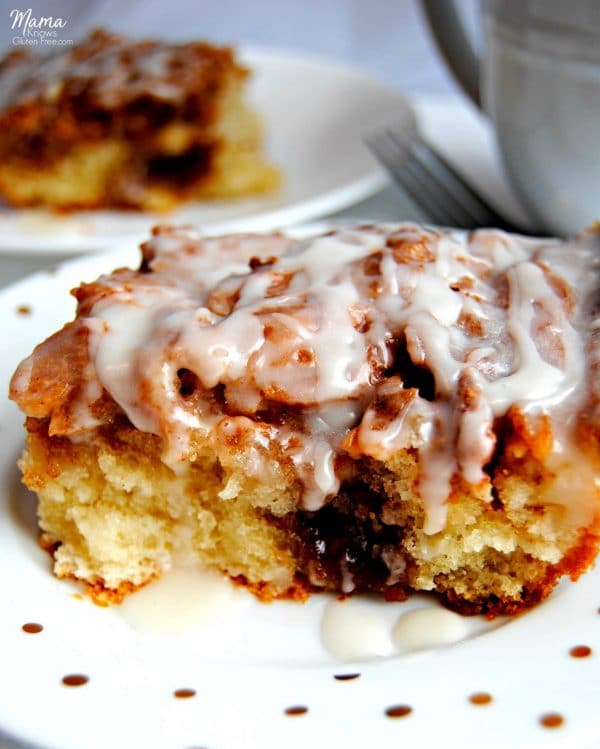 slice of gluten-free cinnamon roll cake on a white plate with another slice and coffee cup in the background