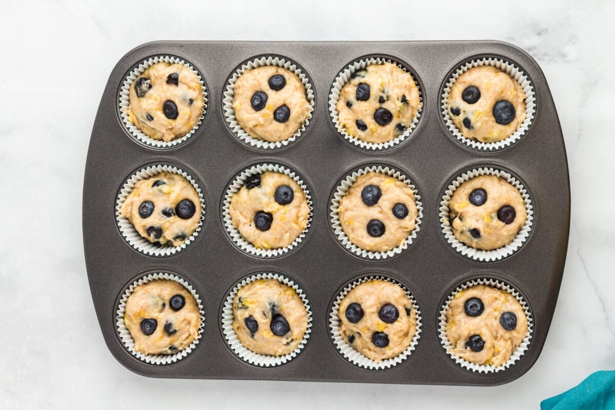 Unbaked gluten-free blueberry banana muffins topped with fresh blueberries in a muffin pan.
