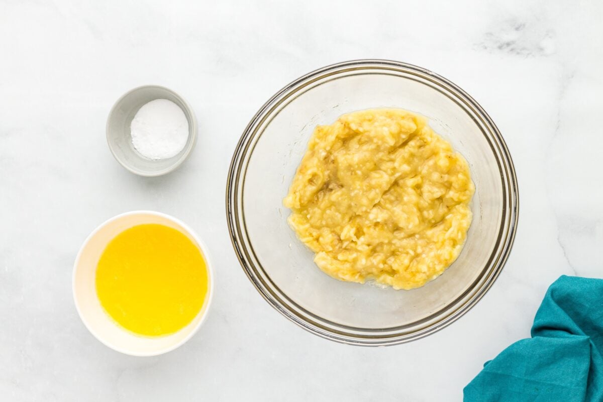 Mashed ripe bananas in a glass bowl with melted butter and baking soda beside the bowl.