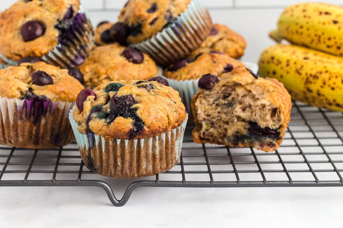Gluten-free blueberry banana muffins cooling on a wire rack with ripe bananas in the background and one muffin sliced open to show the soft, moist interior.