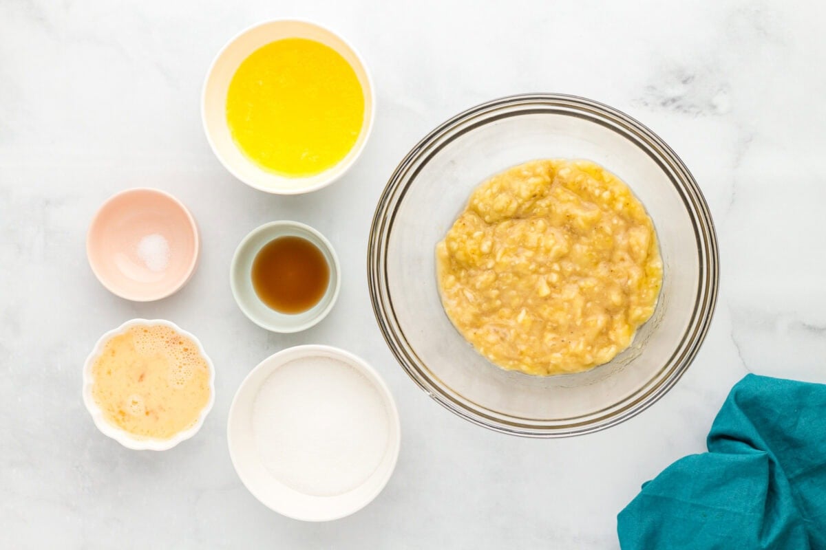 Mashed bananas in a glass bowl with melted butter, sugar, vanilla, salt, and beaten eggs arranged around the bowl.