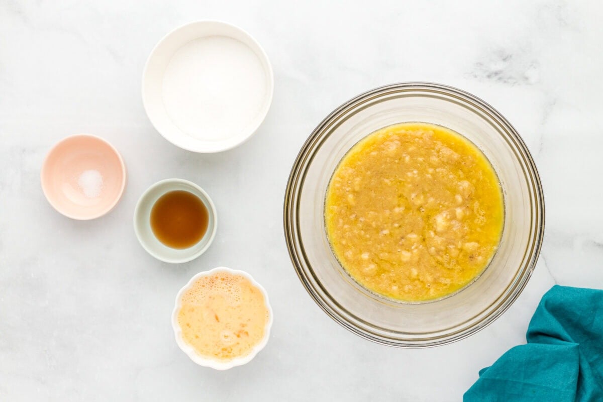 Banana muffin wet ingredients mixed together in a glass bowl with sugar, vanilla, salt, and beaten eggs nearby.
