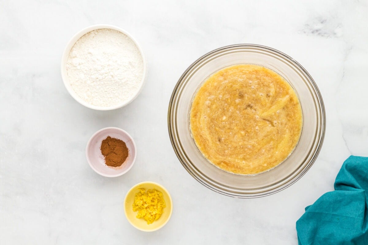 Wet banana muffin mixture in a glass bowl with gluten-free flour, cinnamon, and lemon zest beside the bowl.