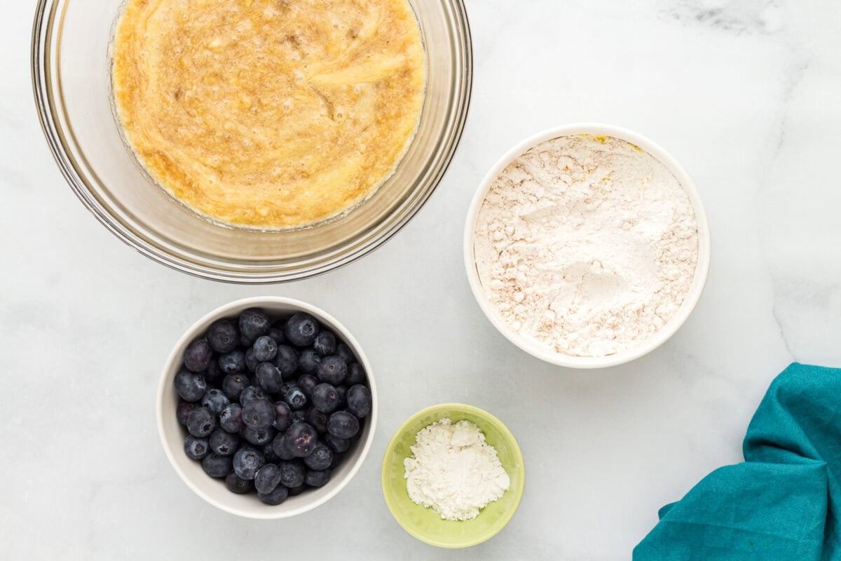 Gluten-free flour and fresh blueberries beside a bowl of banana muffin batter before mixing.