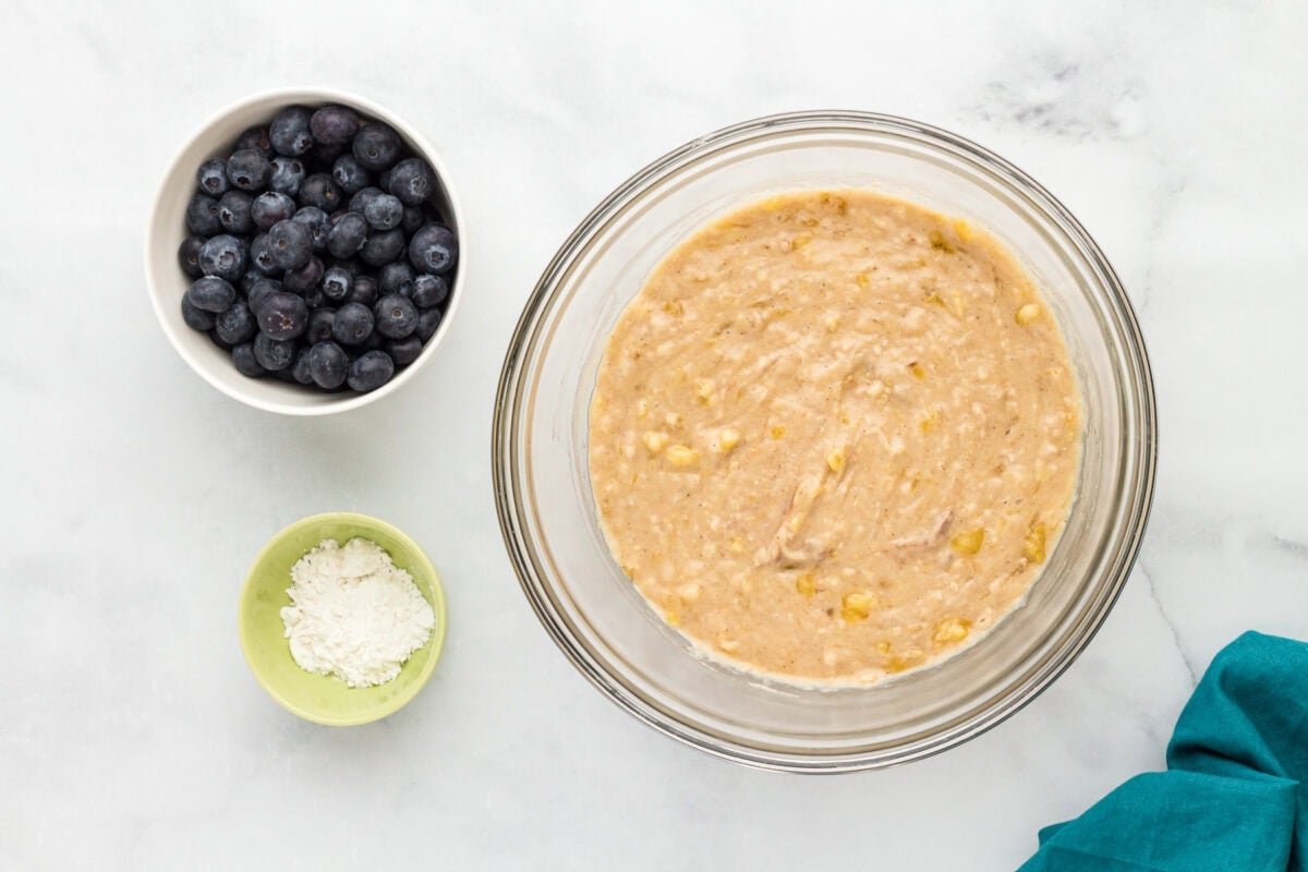 Thick gluten-free blueberry banana muffin batter in a glass bowl with fresh blueberries and reserved flour nearby.
