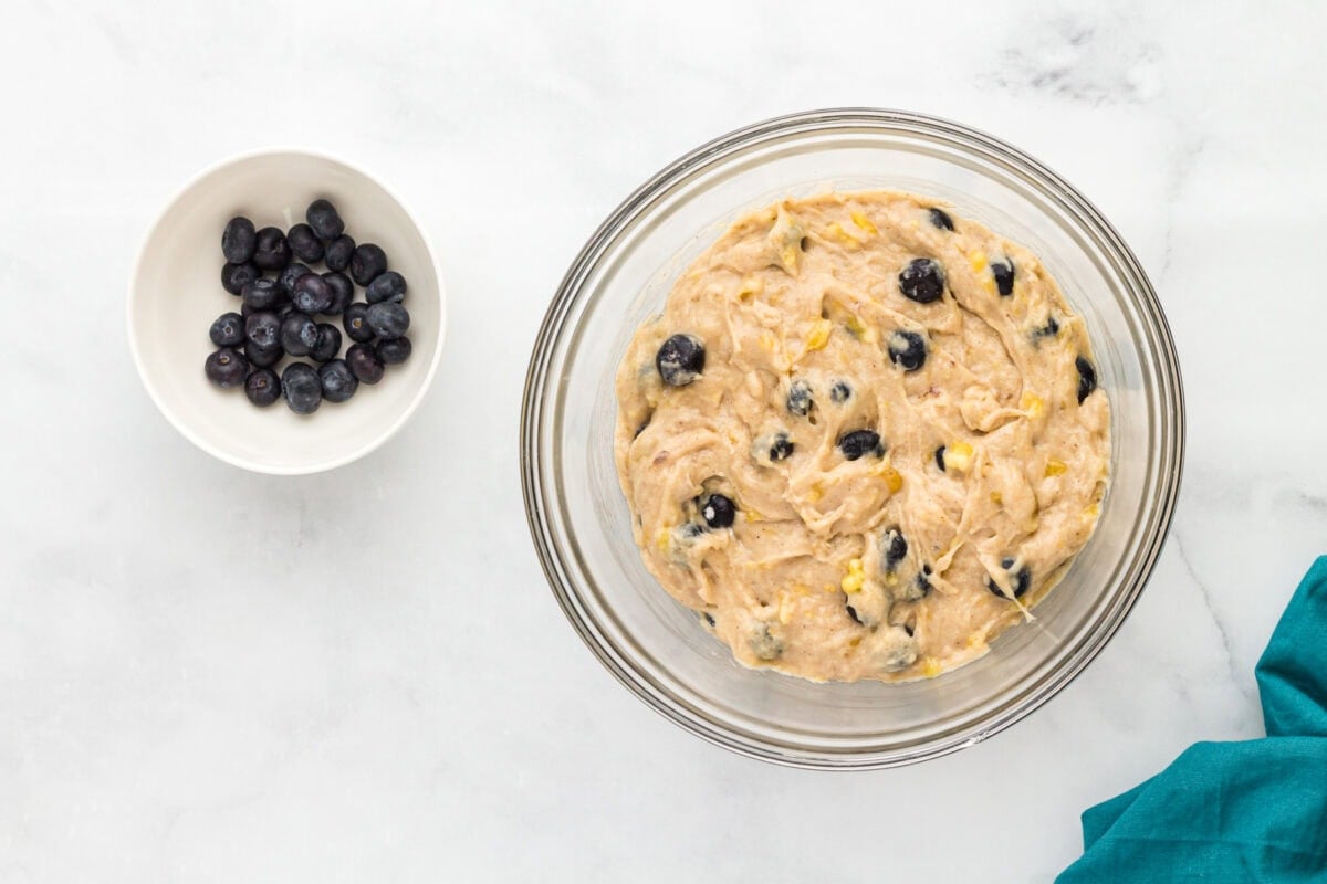Thick gluten-free blueberry banana muffin batter folded with fresh blueberries in a glass mixing bowl.