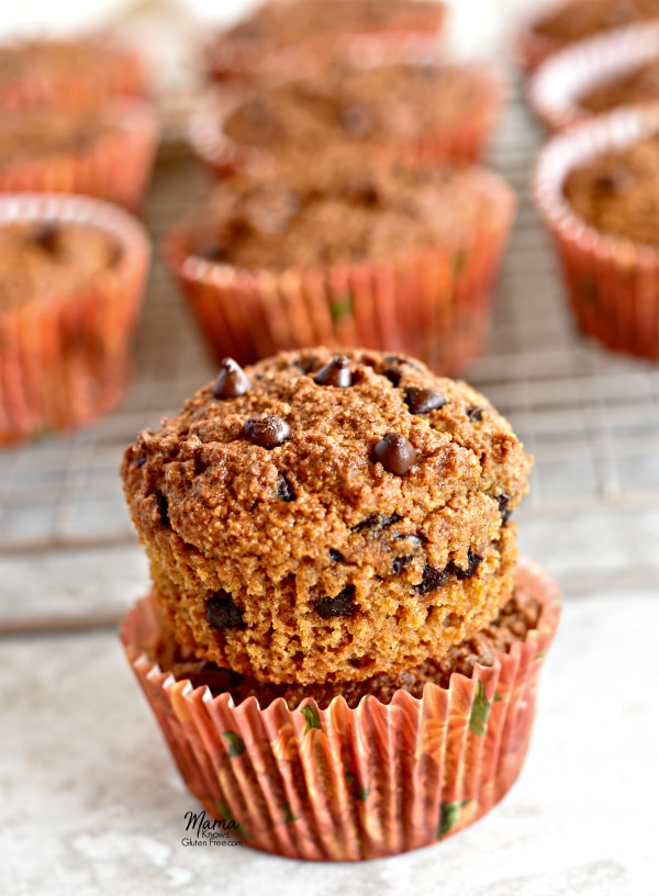Two Paleo pumpkin chocolate chip muffins stacked on top of each other with 9 muffins on a cooling rack behind them.
