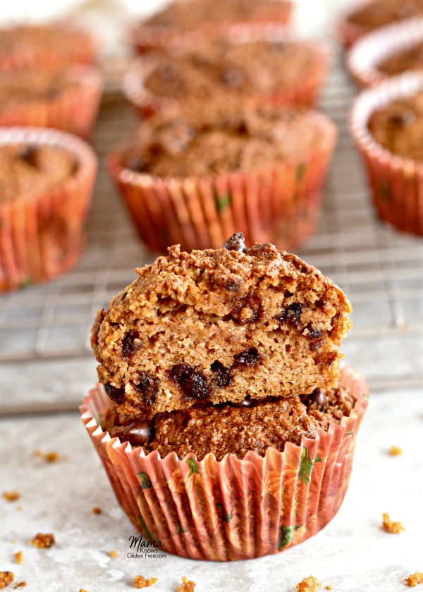 Two Paleo pumpkin chocolate chip muffins stacked on top of each other. One muffin is cut in half to see the texture of the muffin. Six muffins are on a cooling rack in the background.