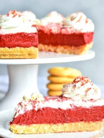 Gluten-Free Red Velvet No-Bake Cheesecake slice on a plate with the cheesecake on a cake stand in the background