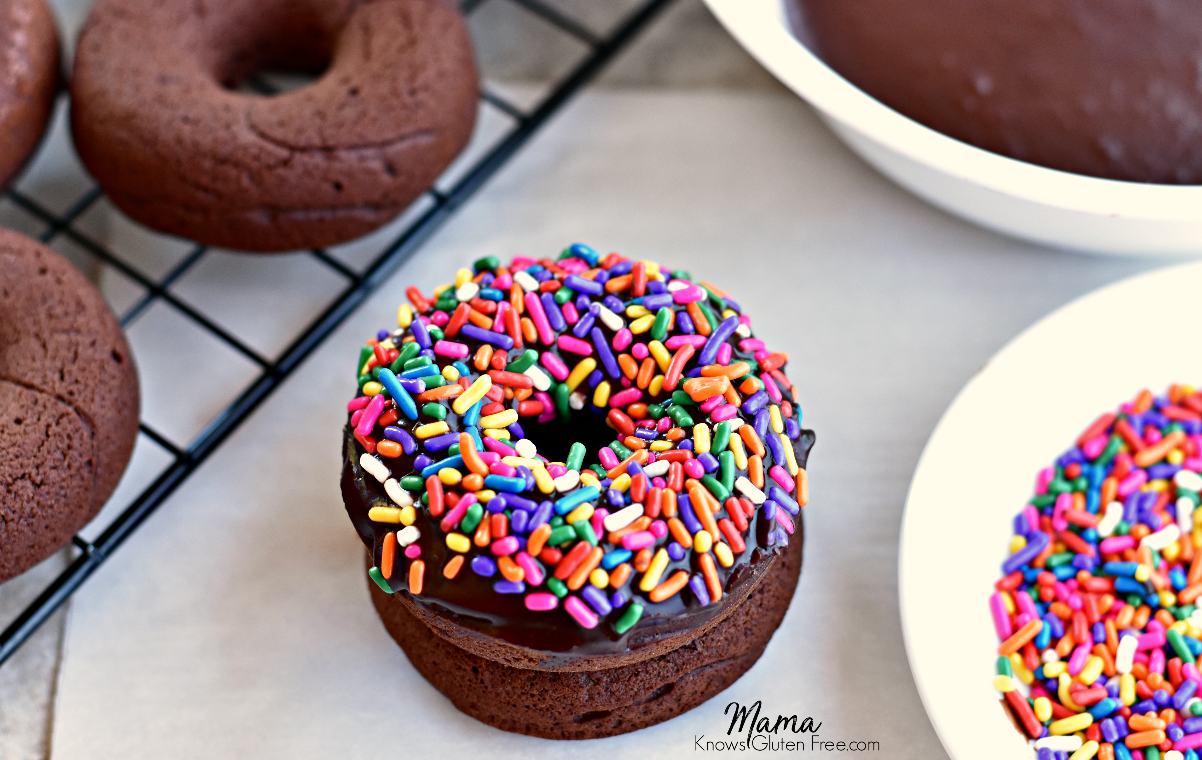 2 gluten-free baked chocolate donuts with sprinkles stacked on top of each other with a bowl of sprinkles, frosting and donuts on a cooling rack