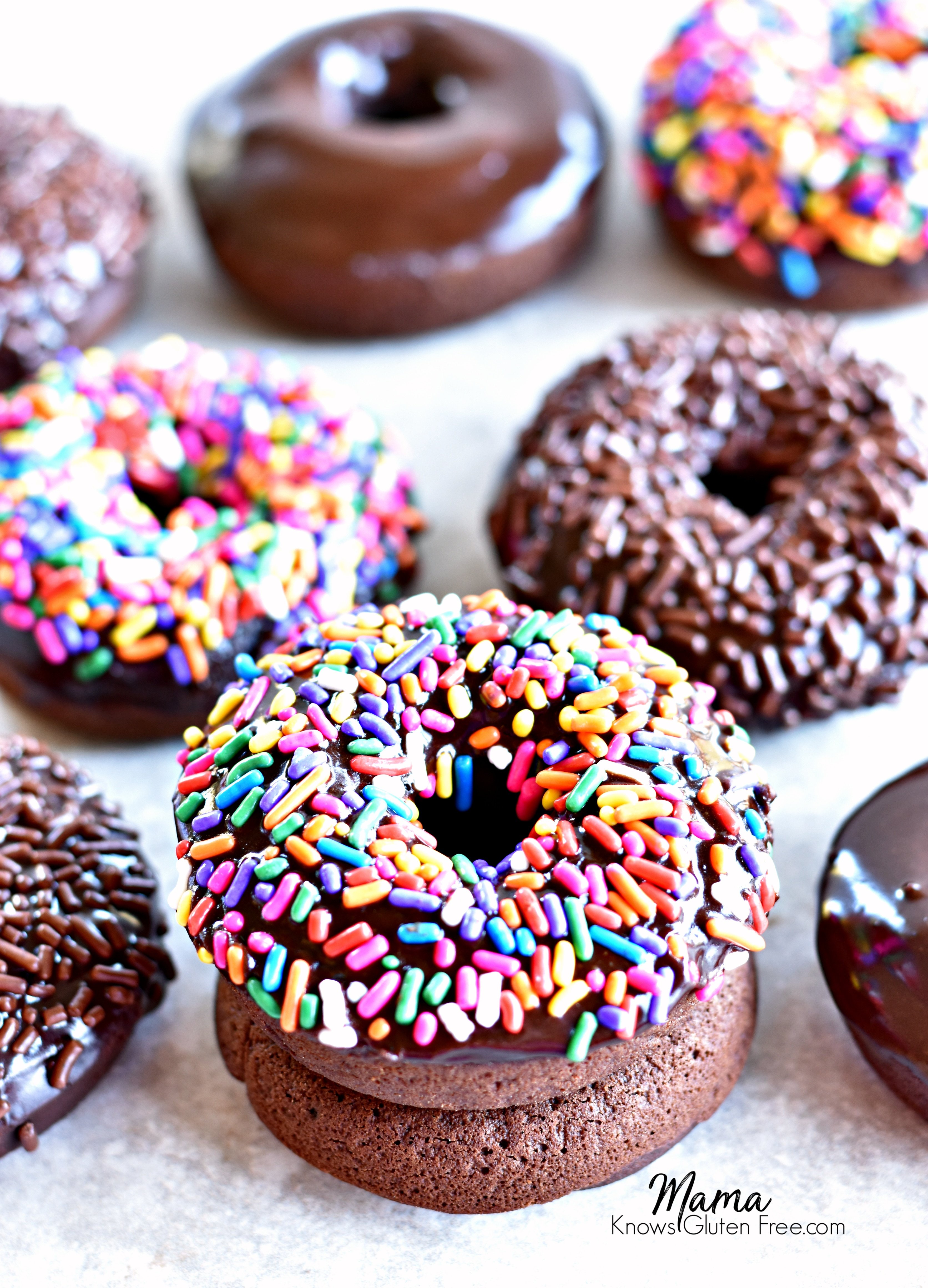 2 gluten-free baked chocolate donuts with sprinkles stacked on top of each other with more donuts in the background