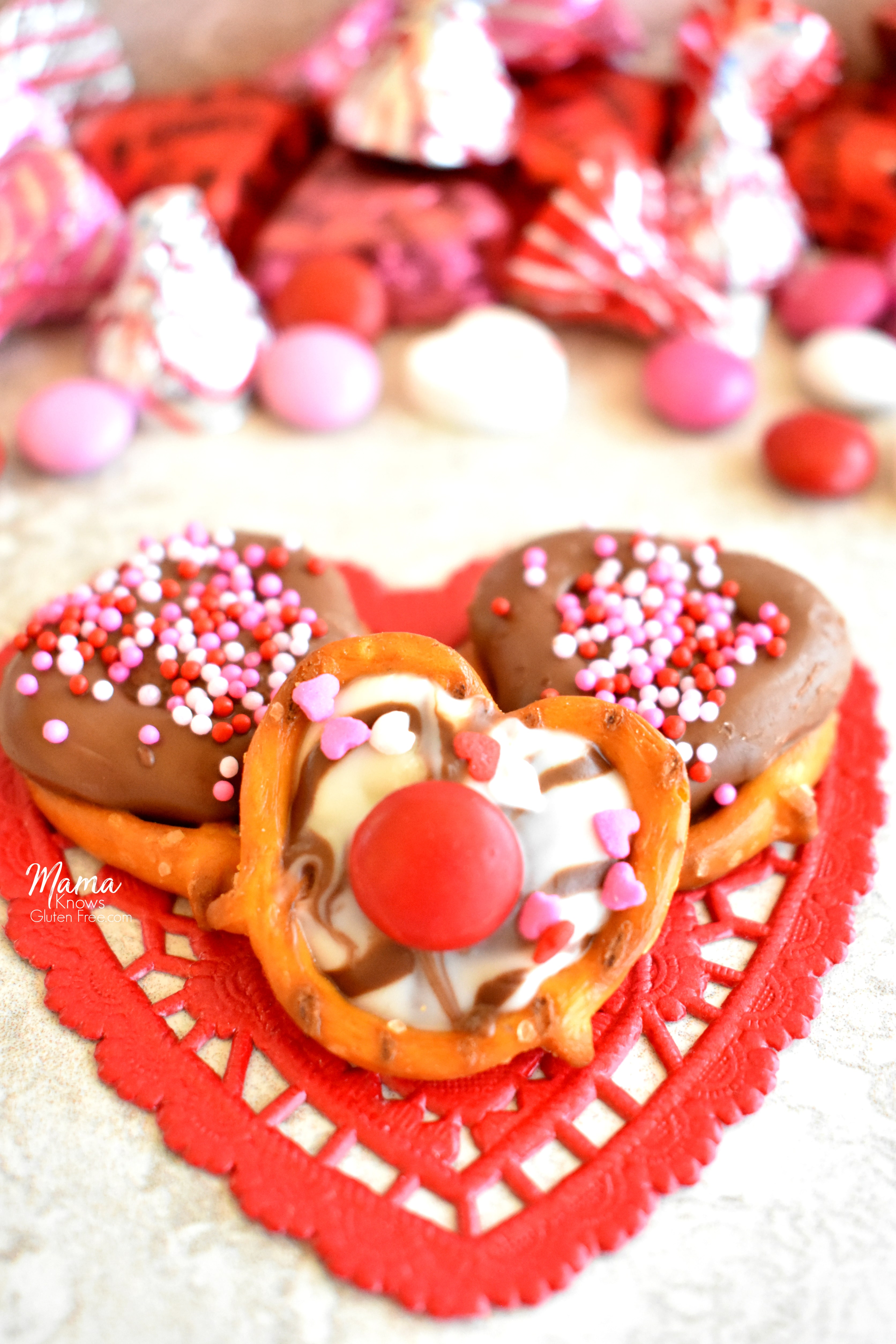 3 gluten-free chocolate covered pretzels on a red heart dollie with candy in the background