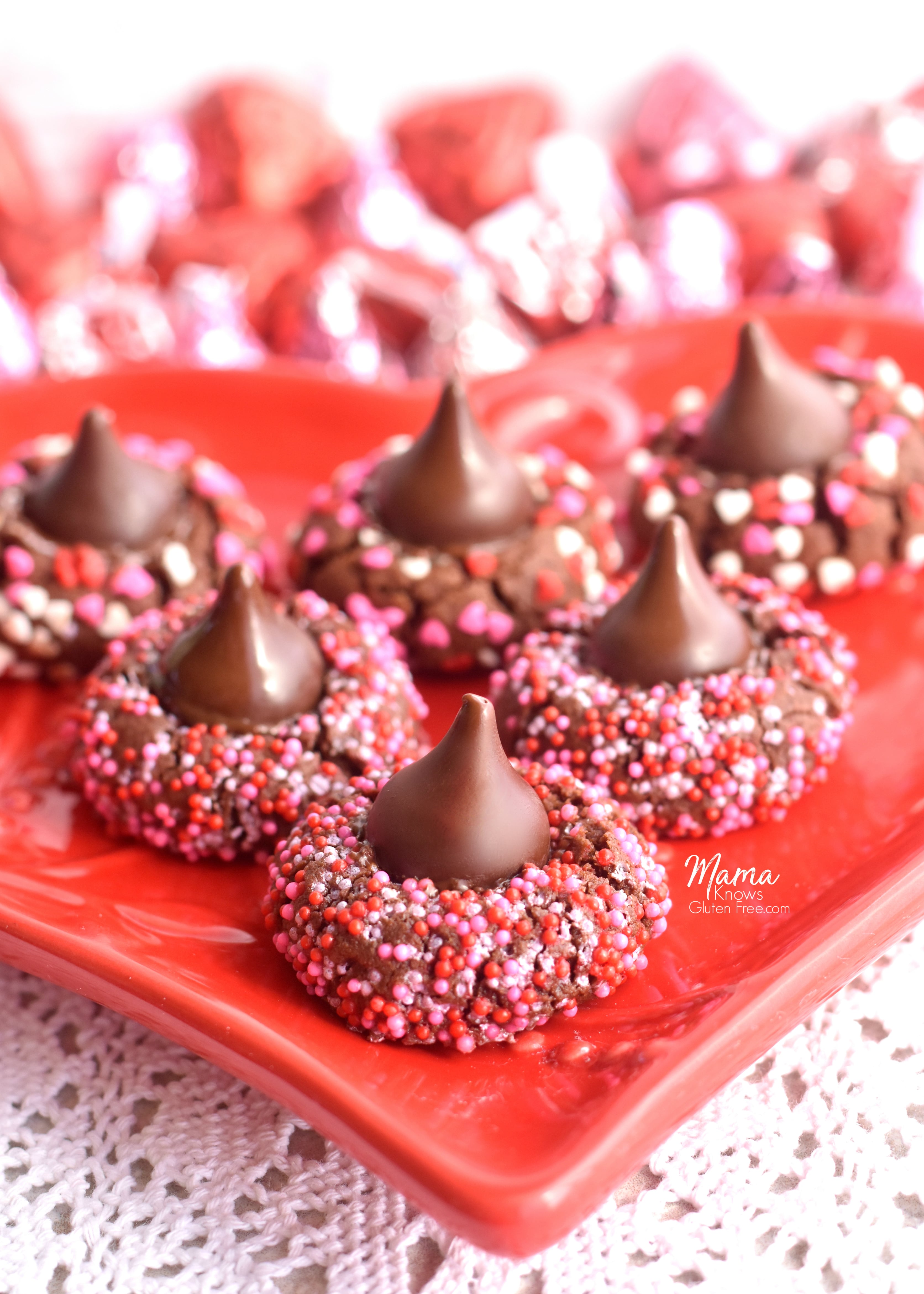 6 gluten-free chocolate kiss cookies on a red heart plate with Hershey kisses in the background