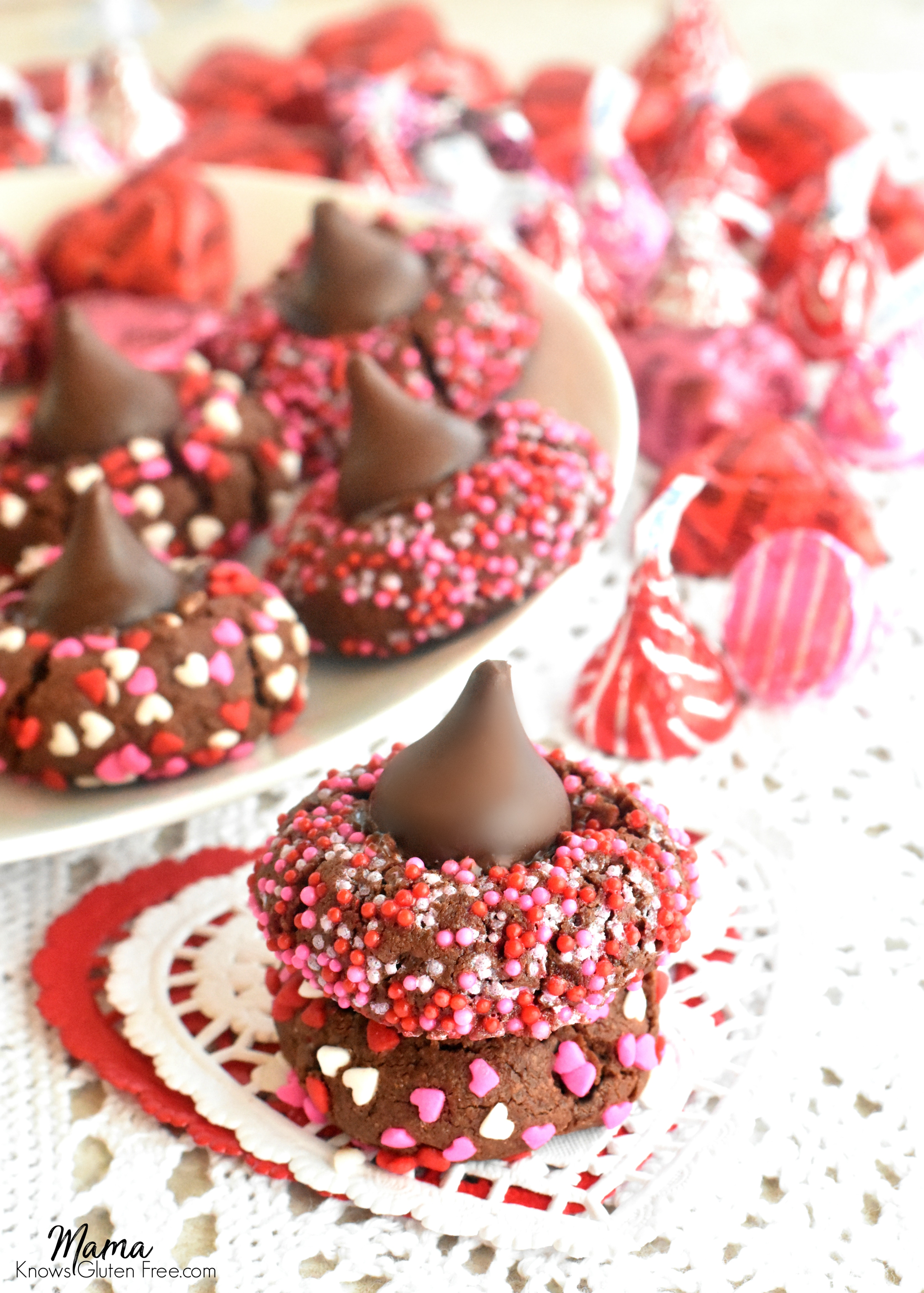 2 gluten-free chocolate kiss cookies stacked on top of each other with a plate of cookies and Hershey kisses in the background