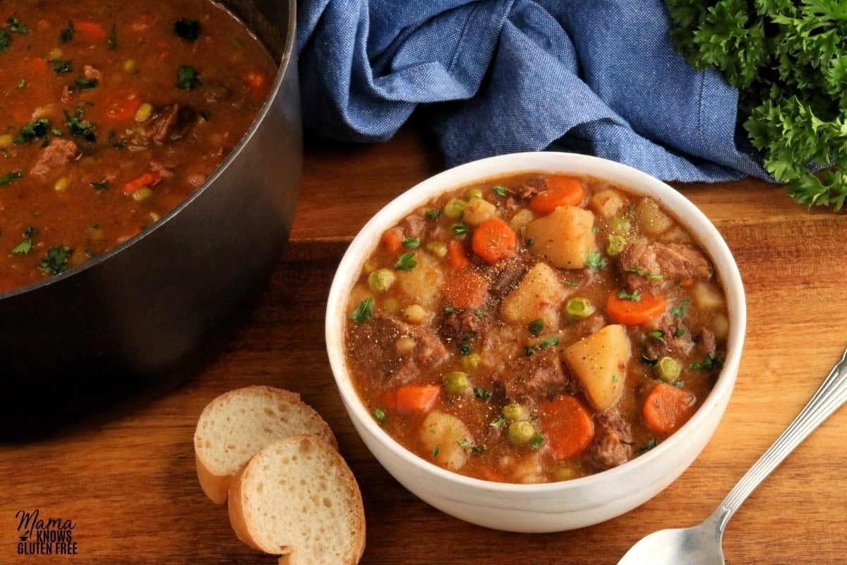 gluten-free beef stew in a white bowl with bread, a spoon and the pot in the background