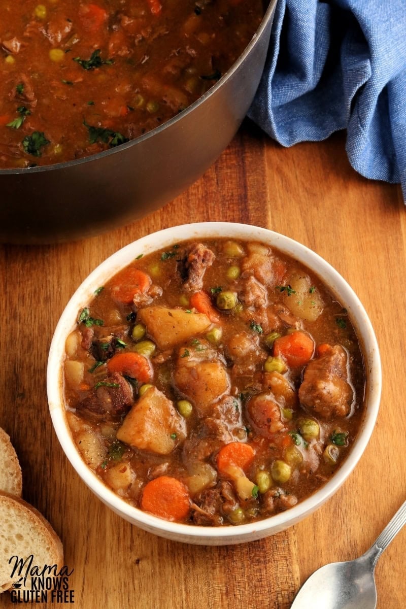 white bowl of gluten-free beef stew, with bread, spoon, and the post and blue napkin in the background 