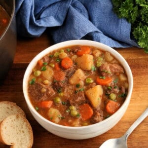 gluten-free beef stew in a white bowl with a spoon, sliced bread, pot, blue napkin, and parsley in the background