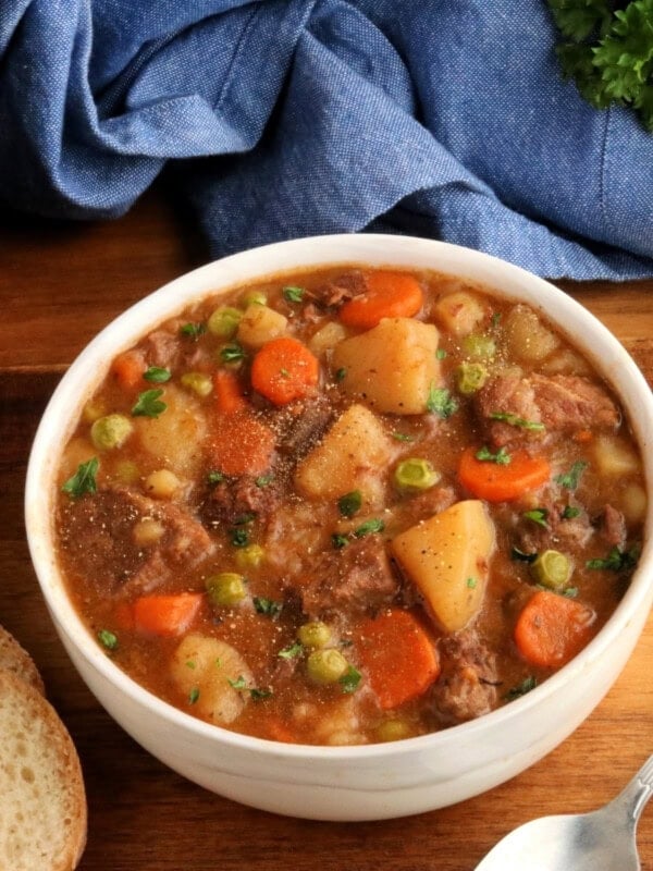 gluten-free beef stew in a white bowl with a spoon, sliced bread, pot, blue napkin, and parsley in the background