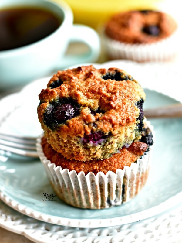 2 Paleo banana blueberry muffins stacked on top of each other on a plate with a fork, coffee cup, muffin and bananas in the background