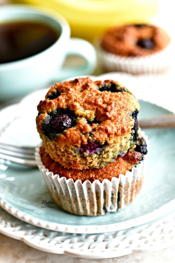 2 Paleo blueberry banana muffins stacked on top of each other on a plate with coffee, a muffins and bananas in the background