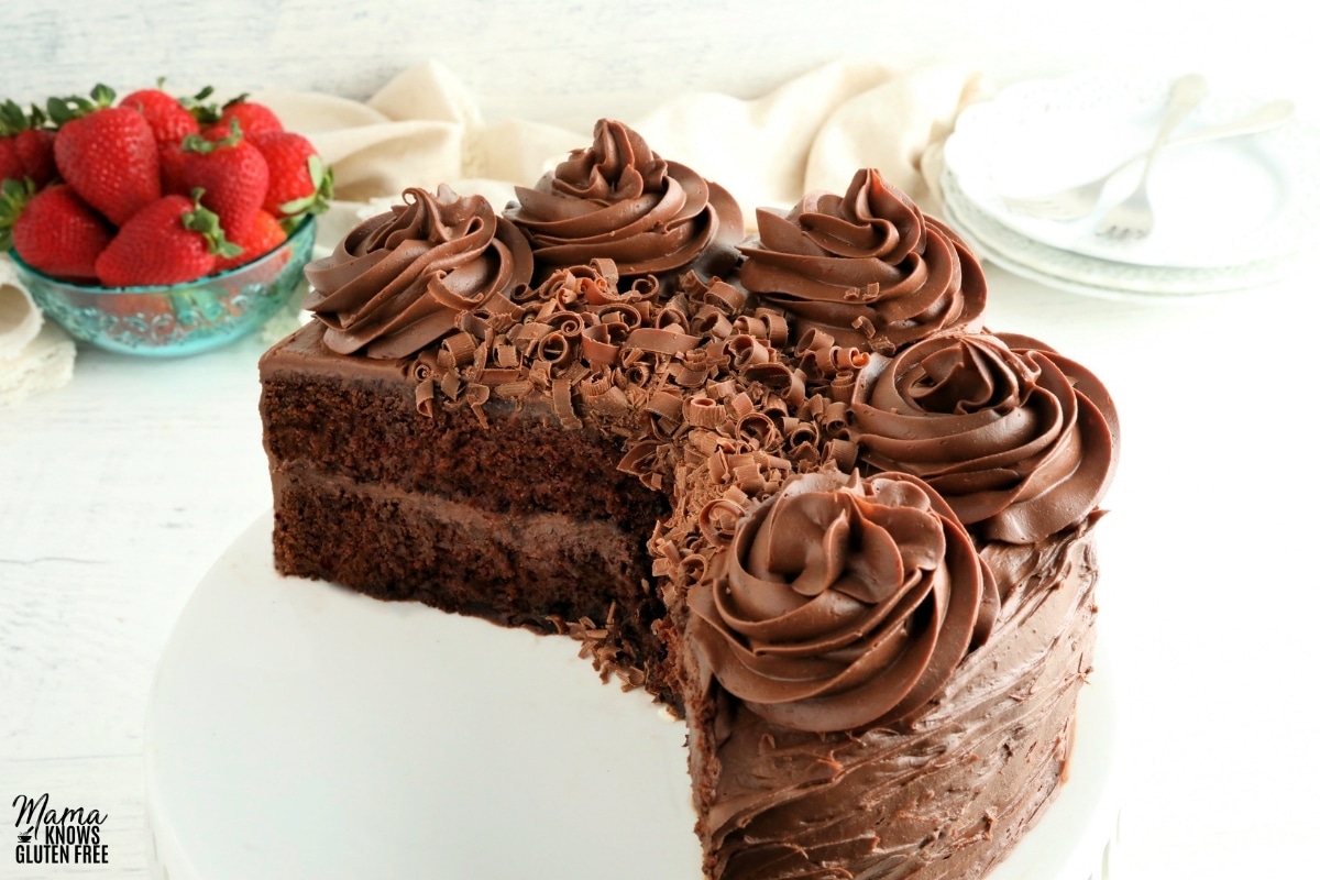 gluten-free chocolate cake that has been sliced on a white cake stand with strawberries n the background