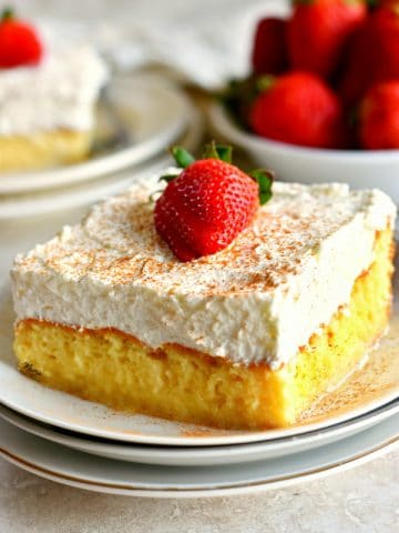 a piece of gluten-free tres leche cake on a plate with more cake and strawberries in the background