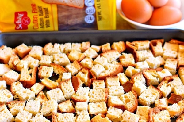 Schar gluten-free bread cut into cubes on a baking sheet with bread and eggs in the background