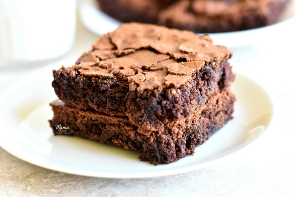 two gluten-free brownies stacked on top of each other with a glass of milk and a plate of brownies in the background