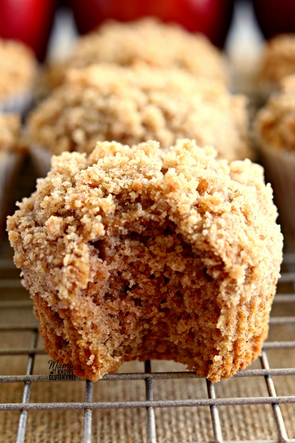 a Gluten-Free Apple Cinnamon Crumb Muffins with a bite out of it to show the texture with more muffins and apples in the background