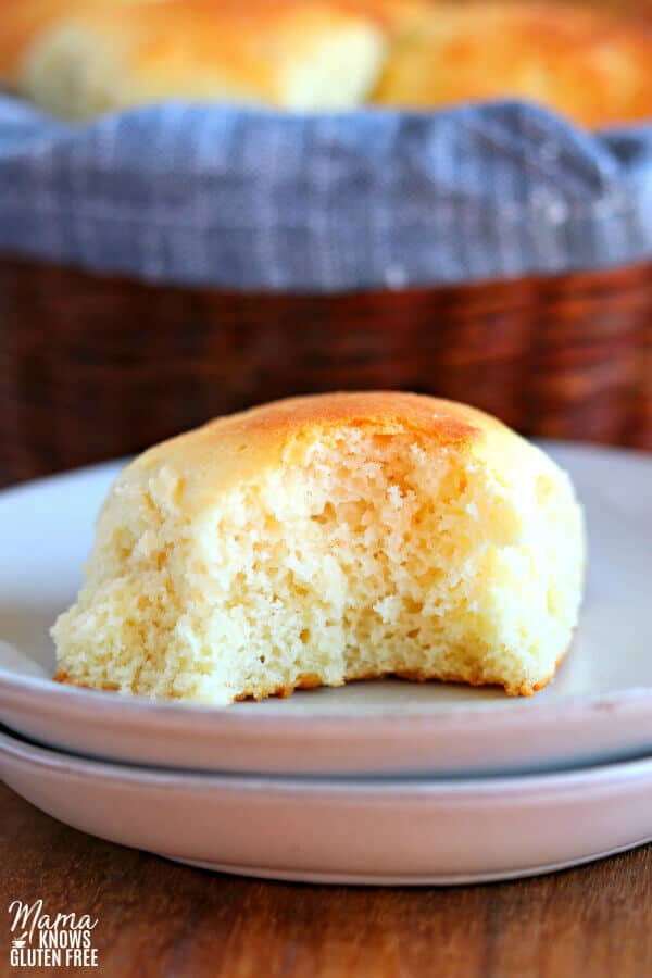 gluten-free dinner roll on a white plate with a bite out of it. Basket of rolls in the background.