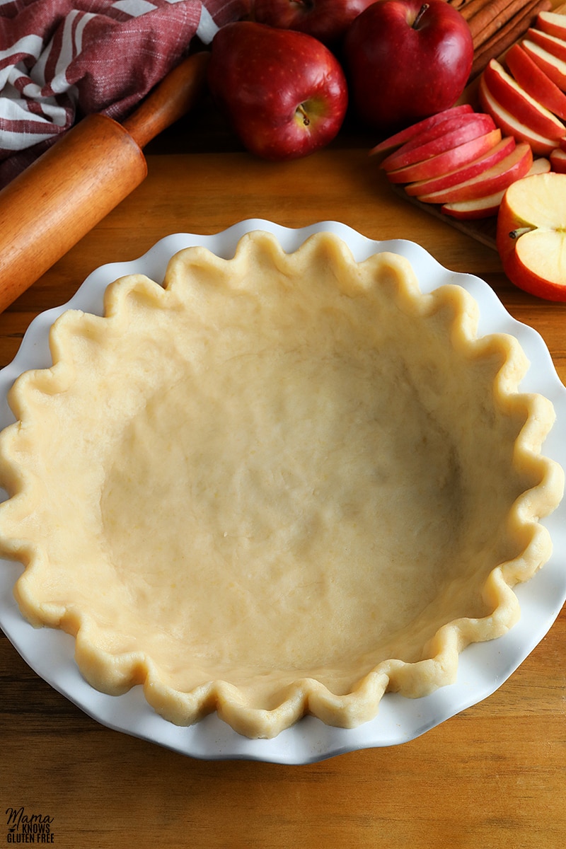 gluten-free pie crust dough in a white pie pan with a pie roller and apples in the background
