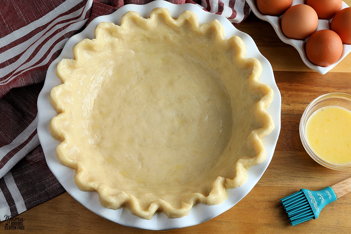 gluten-free pie crust in a white pie pan with egg wash and a pastry brush and eggs.