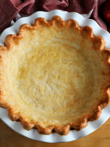 gluten-free pie crust baked in a white pie pan with a red napkin an apples in the background
