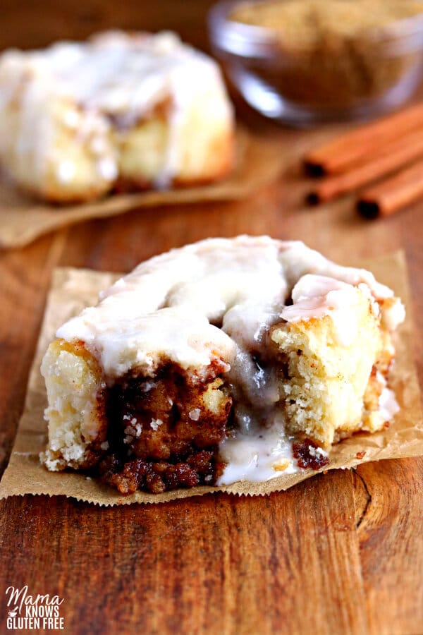 gluten-free cinnamon roll with icing with another cinnamon roll, brown sugar and cinnamon sticks in the background