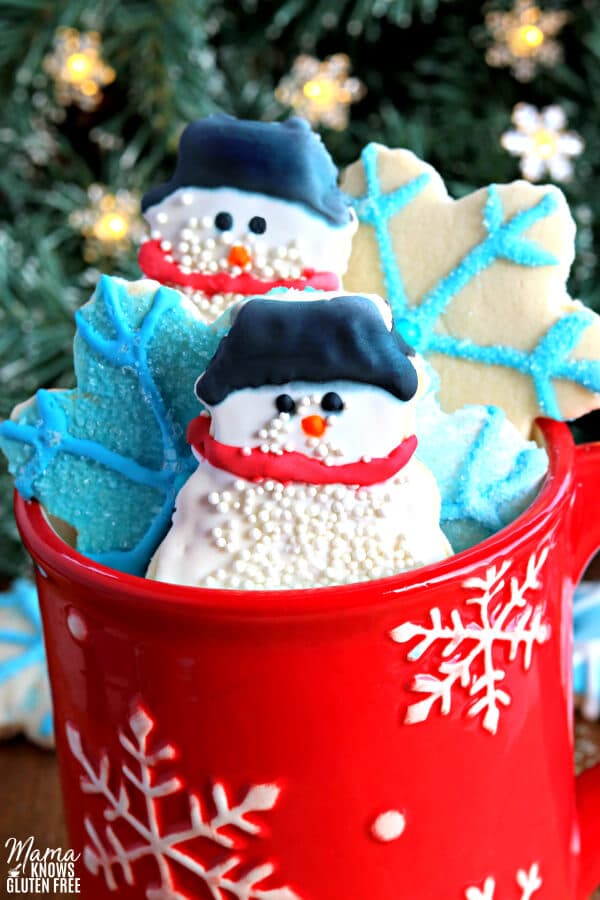 gluten-free sugar cookie cut outs in snow man and snowflakes in a red cup with a Christmas tree in the background