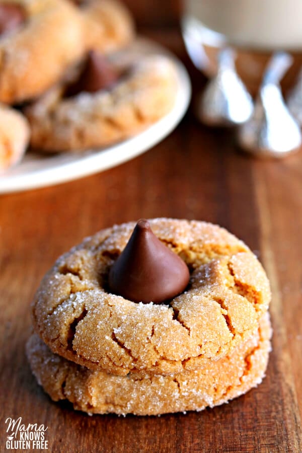 2 gluten-free peanut butter blossom cookies stacked on to of each other with cookies on a plate, Hershey's kisses and glass of milk in the background