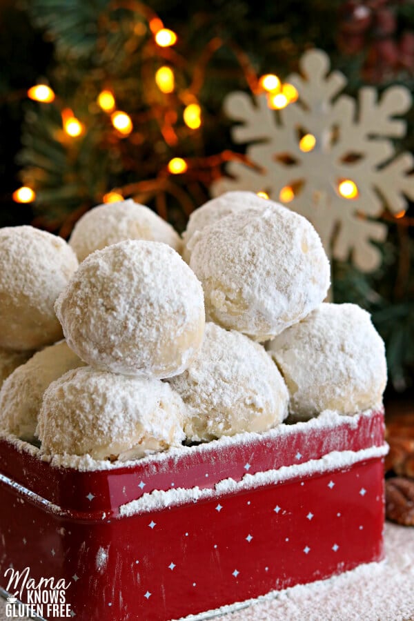 gluten-free snowball cookies in a red cookie tin with a snowflake and lighted tree in the background