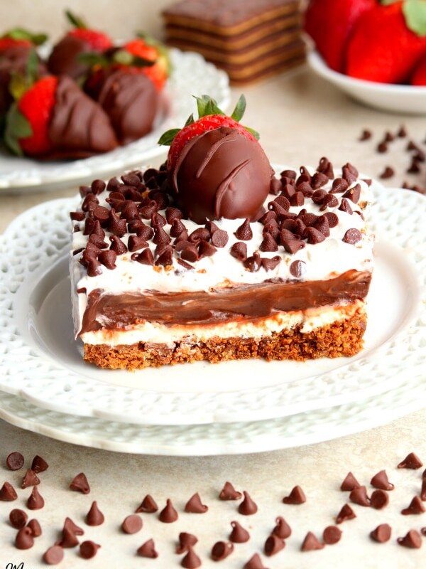 a slice of chocolate lasagna with chocolate covered strawberries, cookies, strawberries and chocolate chips in the background