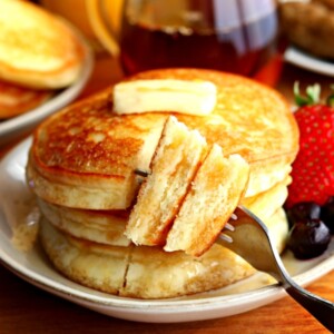 a stack of 3 gluten-free pancakes cut with a bite on a fork with butter, syrup, blueberries and strawberry on a plate with syrup, sausage, orange juice and a plate of pancakes in the background