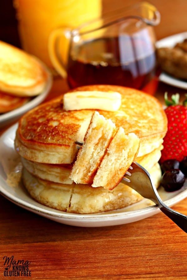 a stack of 3 gluten-free pancakes cut with a bite on a fork with butter, syrup, blueberries and strawberry on a plate with syrup, sausage, orange juice and a plate of pancakes in the background