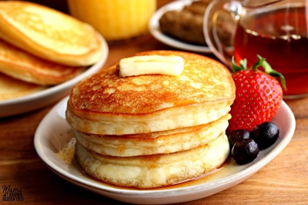 stack of three pancakes with a strawberry and blueberries on a plate with sausage, syrup, orange juice and a plate of pancakes in the background