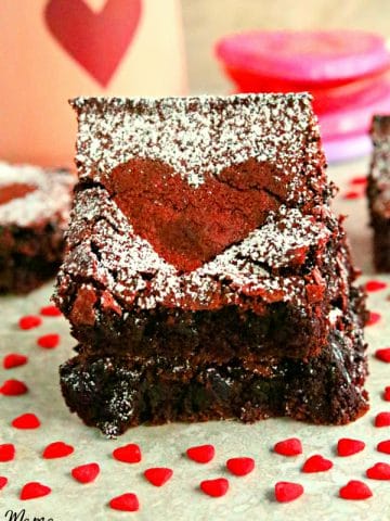 2 gluten-free red velvet brownies stacked on top of each other with more bownies, a heart cup and heart candy in the background