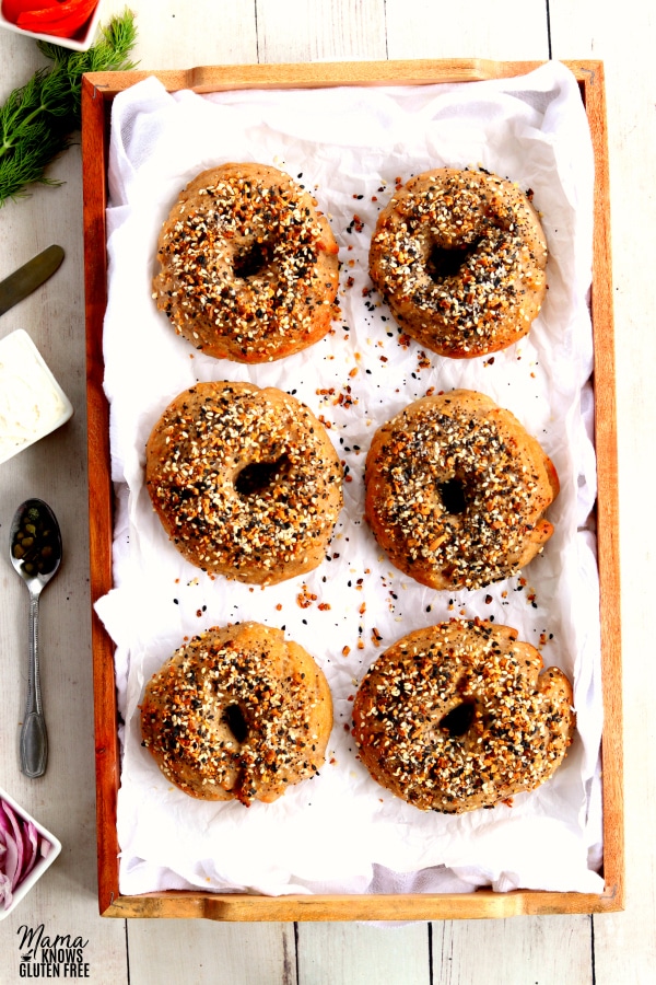 6 gluten-free bagels on a wood try with locks, dill, cream cheese, a spoon with capers and chopped onion son the side