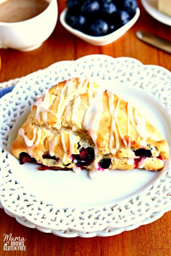 a white plate with a blueberry scone on it with a cup of coffee and blueberries and butter in the background